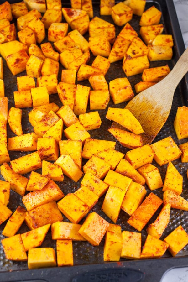 Uncooked butternut squash chunks tossed in olive oil and spices on baking sheet.