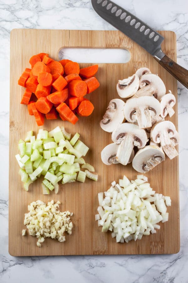 Minced garlic, onions, celery, carrots, and mushrooms on wooden cutting board.