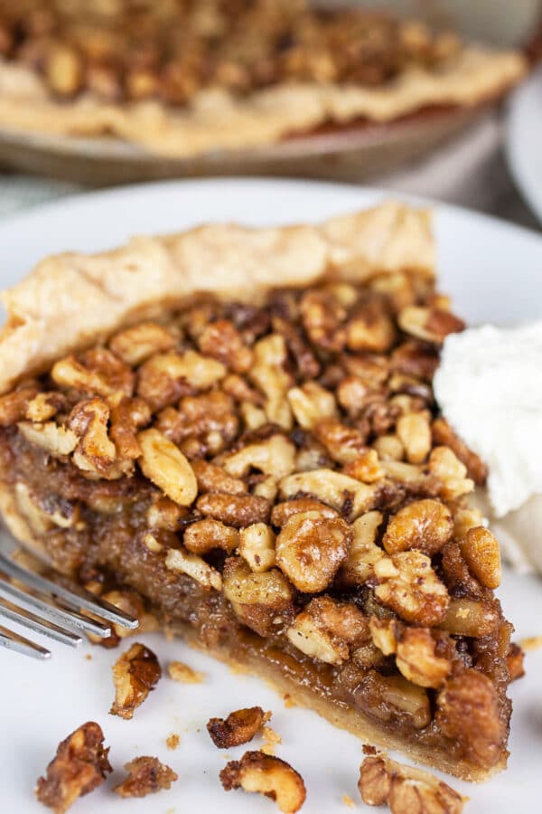 This Maple Bourbon Walnut Pie puts a twist on the traditional pecan pie. It's sweetened with maple syrup and kicked up with a shot of bourbon. This homemade pie is one of my favorite dessert recipes. I will take this maple bourbon walnut pie over pecan any day! #pie #maple #bourbon #walnut #thanksgiving Piece of maple bourbon walnut pie on white plate.