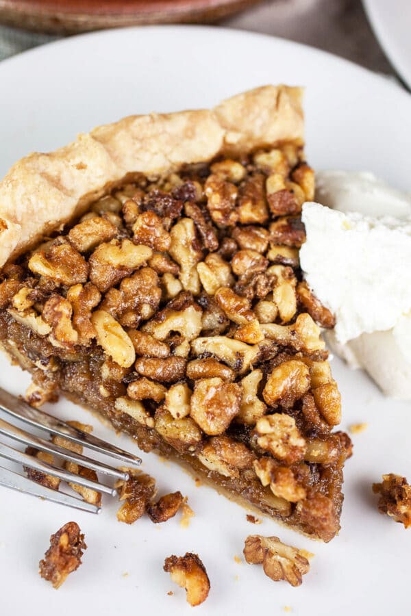 This Maple Bourbon Walnut Pie puts a twist on the traditional pecan pie. It's sweetened with maple syrup and kicked up with a shot of bourbon. This homemade pie is one of my favorite dessert recipes. I will take this maple bourbon walnut pie over pecan any day! #pie #maple #bourbon #walnut #thanksgiving Piece of toasted walnut pie with whipped cream on small white plate with fork.