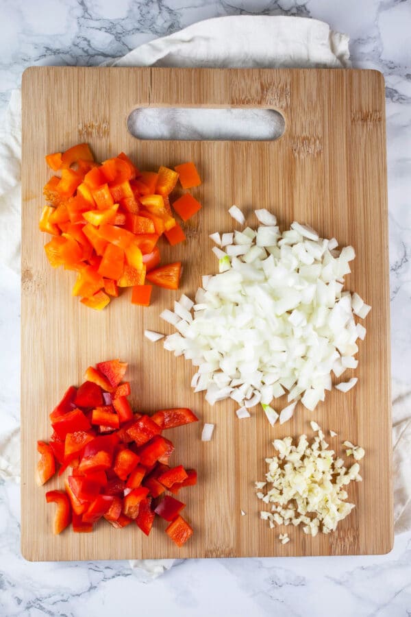 Minced garlic, onions, red peppers, and orange peppers on wooden cutting board.