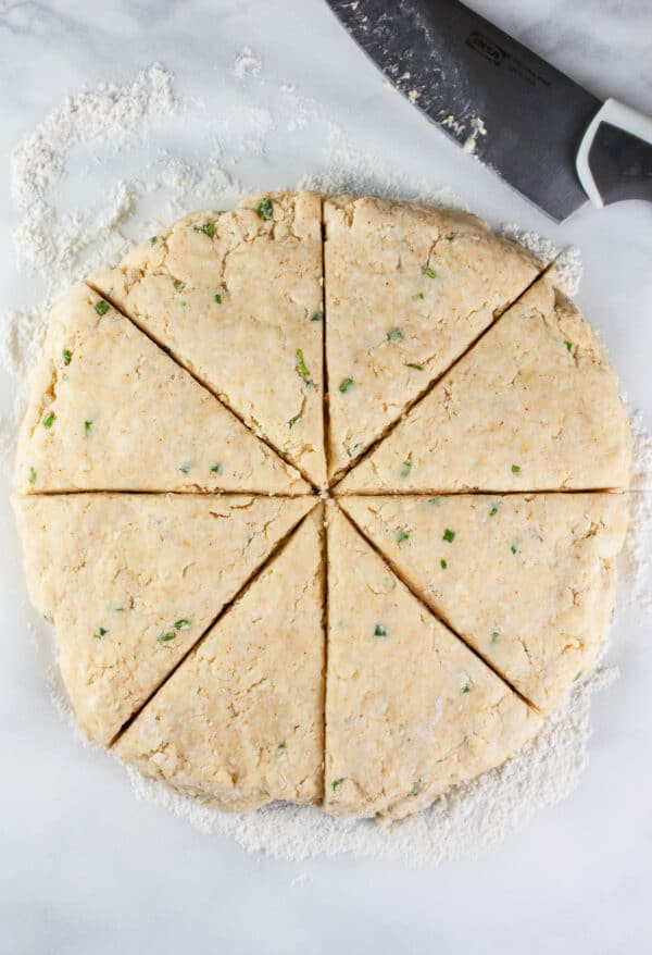 Round disc of scone dough cut into wedges on white surface.