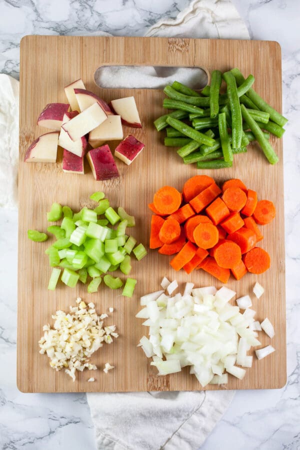 Minced garlic, onions, celery, carrots, red potatoes, and green beans on wooden cutting board.