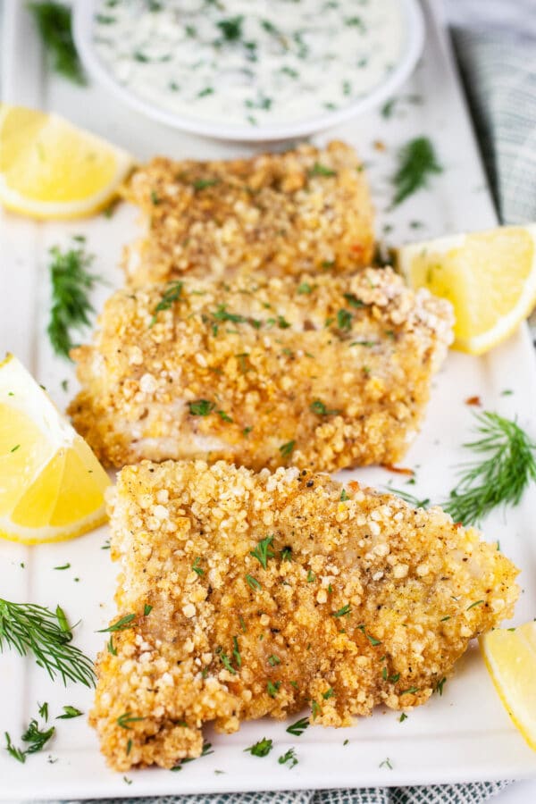 Breaded walleye fingers on white platter with small bowl of tartar sauce, lemon wedges, and fresh dill.