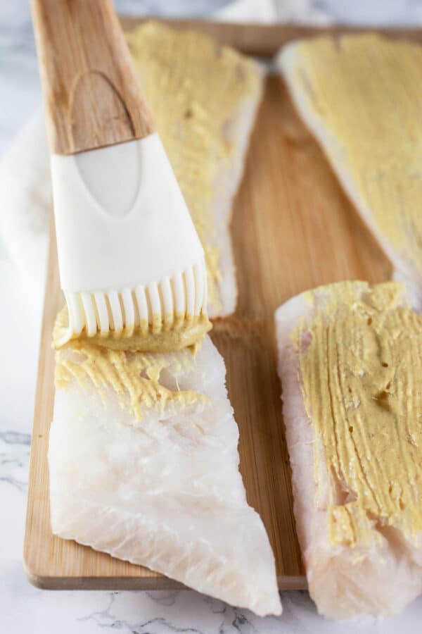 Dijon mustard brushed onto cod fillets on wooden cutting board.