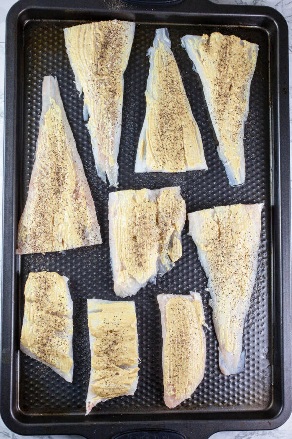 Uncooked cod fillets with Dijon mustard on baking sheet.