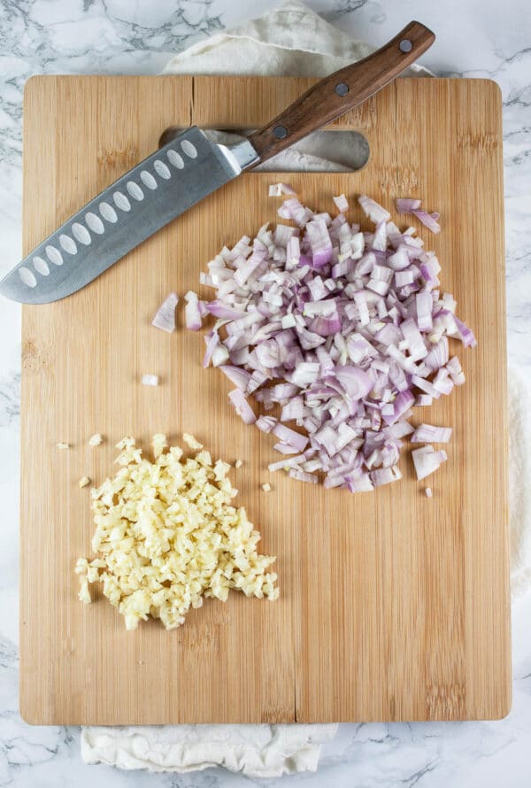 Minced garlic and shallots on wooden cutting board with knife.