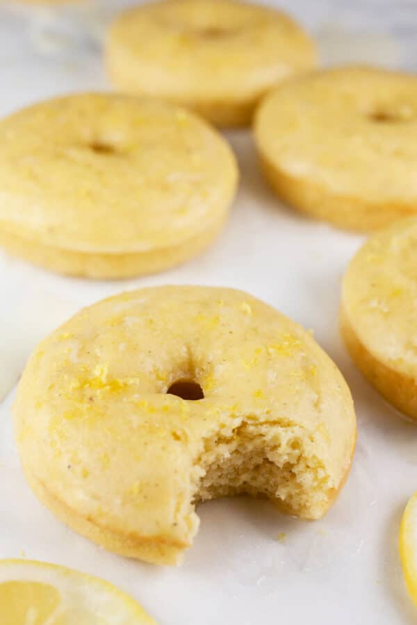 Baked lemon doughnuts on white surface, one with bite taken out.