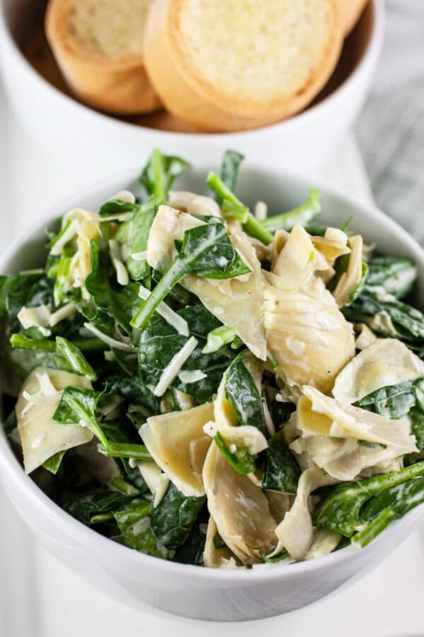 This Lemony Spinach Artichoke Dip is a lightened up version of the classic! This recipe is so easy to make and uses fresh spinach and artichokes. It's light and lemony and focuses on the flavor of the ingredients. This simple appetizer is perfect for those who want a healthy version of this iconic dip. Makes enough to feed a crowd so it's great for picnics or parties! #spinachartichokedip #spinach #artichoke #dips #appetizers #recipes #healthy Cold spinach artichoke dip in small white bowl with crostini on serving platter.
