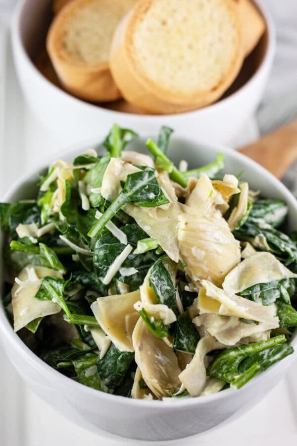 Spinach artichoke dip with Parmesan cheese and crostini in small white bowls.