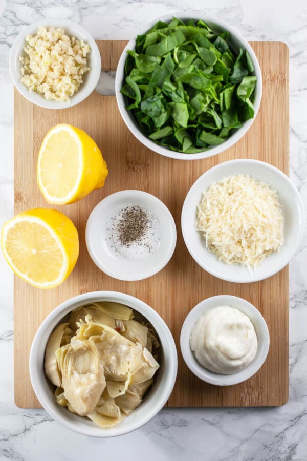 Marinated artichokes, mayo, Parmesan cheese, salt, pepper, lemons, garlic, and fresh spinach on wooden cutting board.