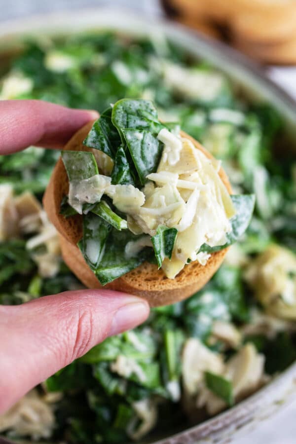 Fingers holding spinach artichoke dip crostini in front of ceramic bowl.
