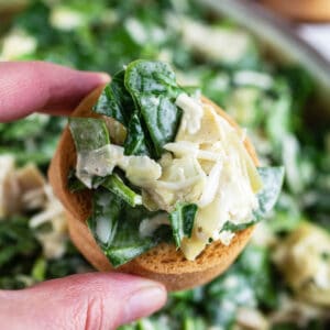 Fingers holding spinach artichoke dip on crostini in front of ceramic bowl.