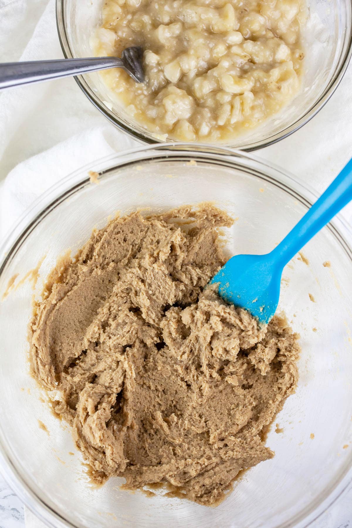 Butter and brown sugar creamed in large glass bowl next to small glass bowl of mashed bananas.