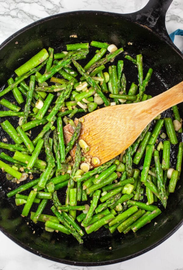 Garlic, green onions, and asparagus sautéed in cast iron skillet with wooden spatula.