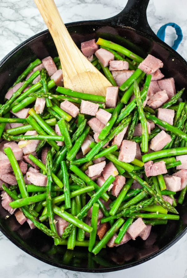 Diced ham and asparagus sautéed in cast iron skillet with wooden spoon.