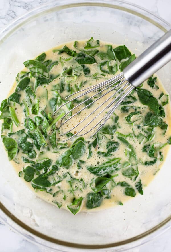 Egg mixture with chopped spinach in large glass mixing bowl with whisk.