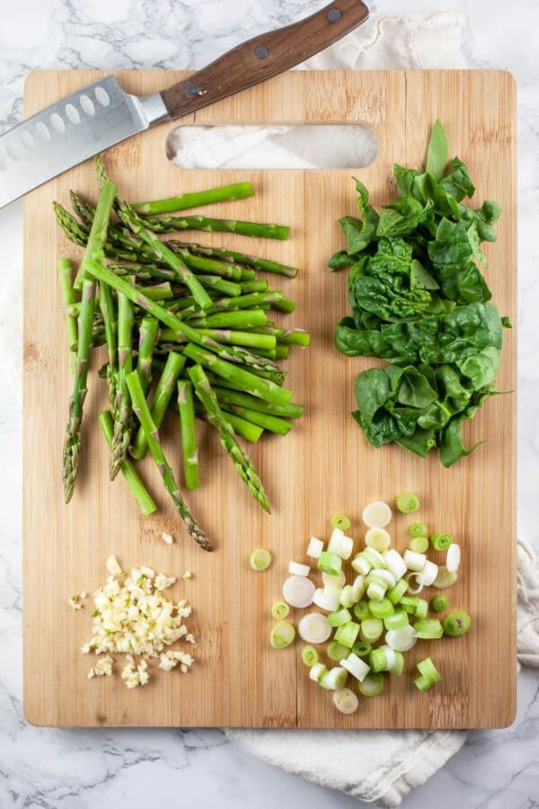 Minced garlic and green onions and chopped asparagus and spinach on wooden cutting board with knife.