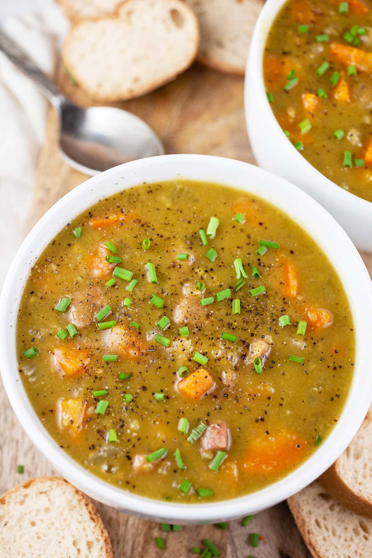 Split pea and ham soup in white bowls with slices of bread on wooden serving board.