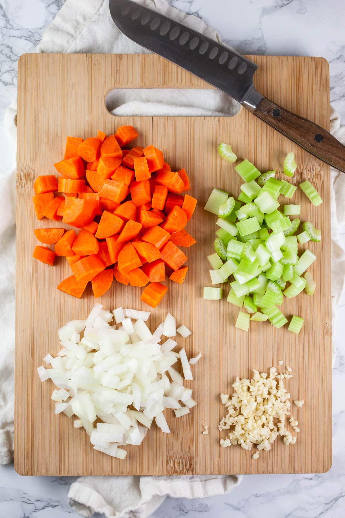 Minced garlic and onions and chopped celery and carrots on wooden cutting board with knife.