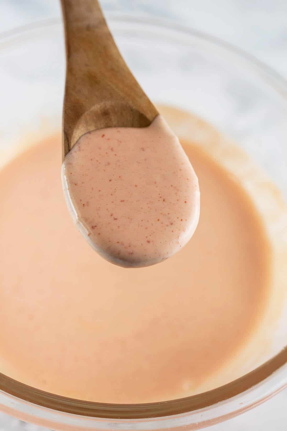 Spoonful of Sriracha mayo on wooden spoon lifted from small glass bowl.