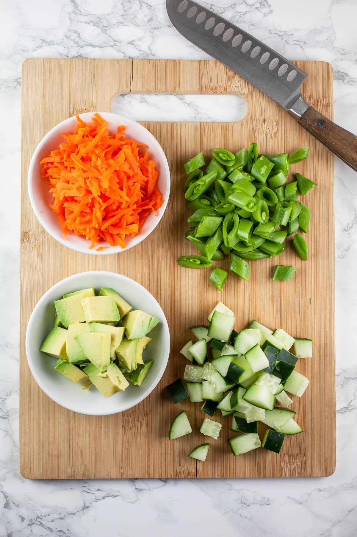 Diced avocado, cucumbers, snap peas, and grated carrots on wooden cutting board with knife.