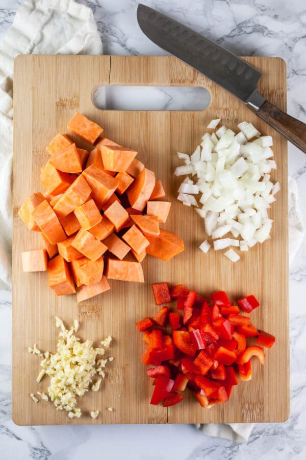 Minced garlic, onions, red bell peppers, and sweet potatoes on wooden cutting board with knife.