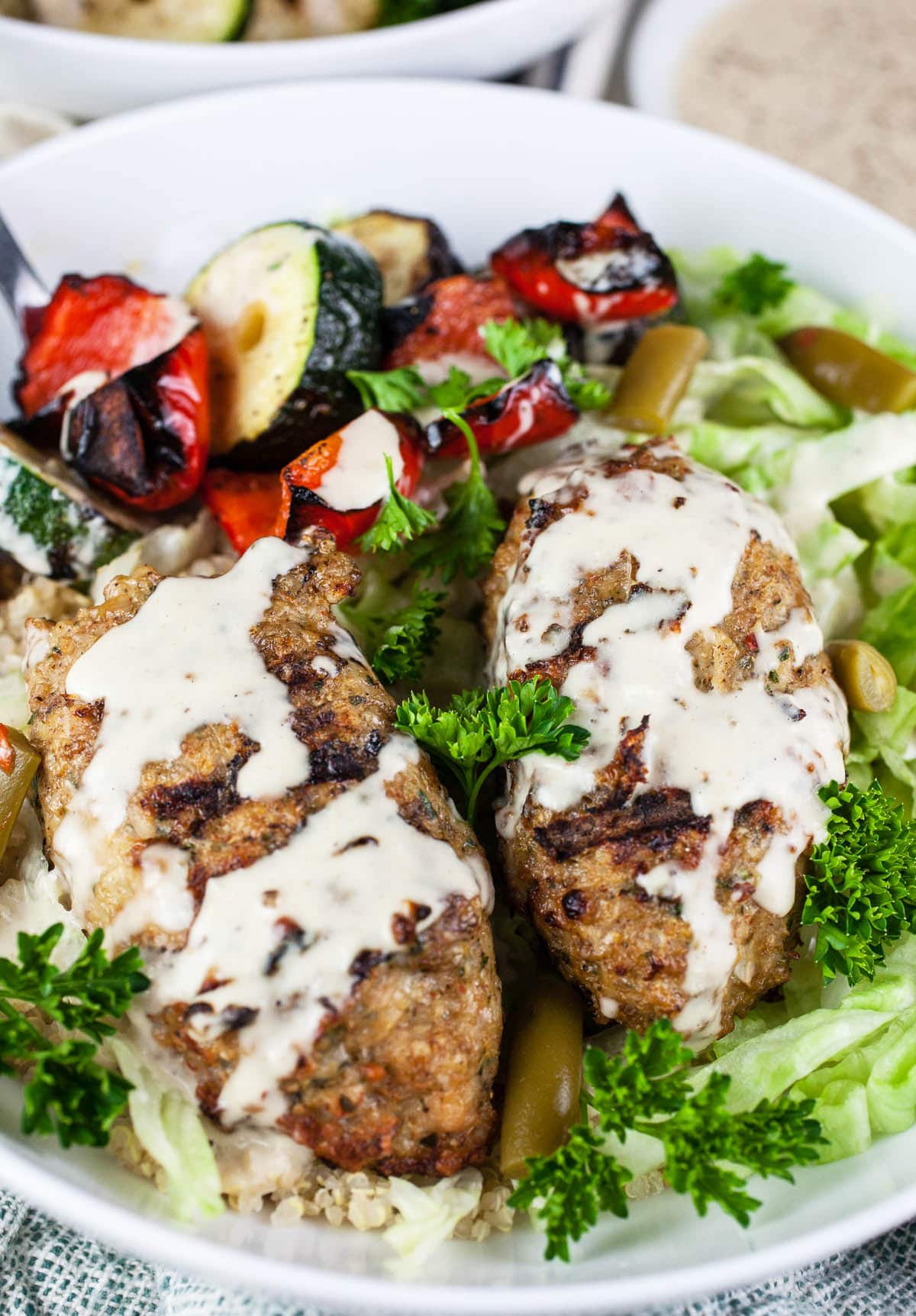 Chicken kofta kebabs with grilled vegetables and tahini sauce in white bowls.