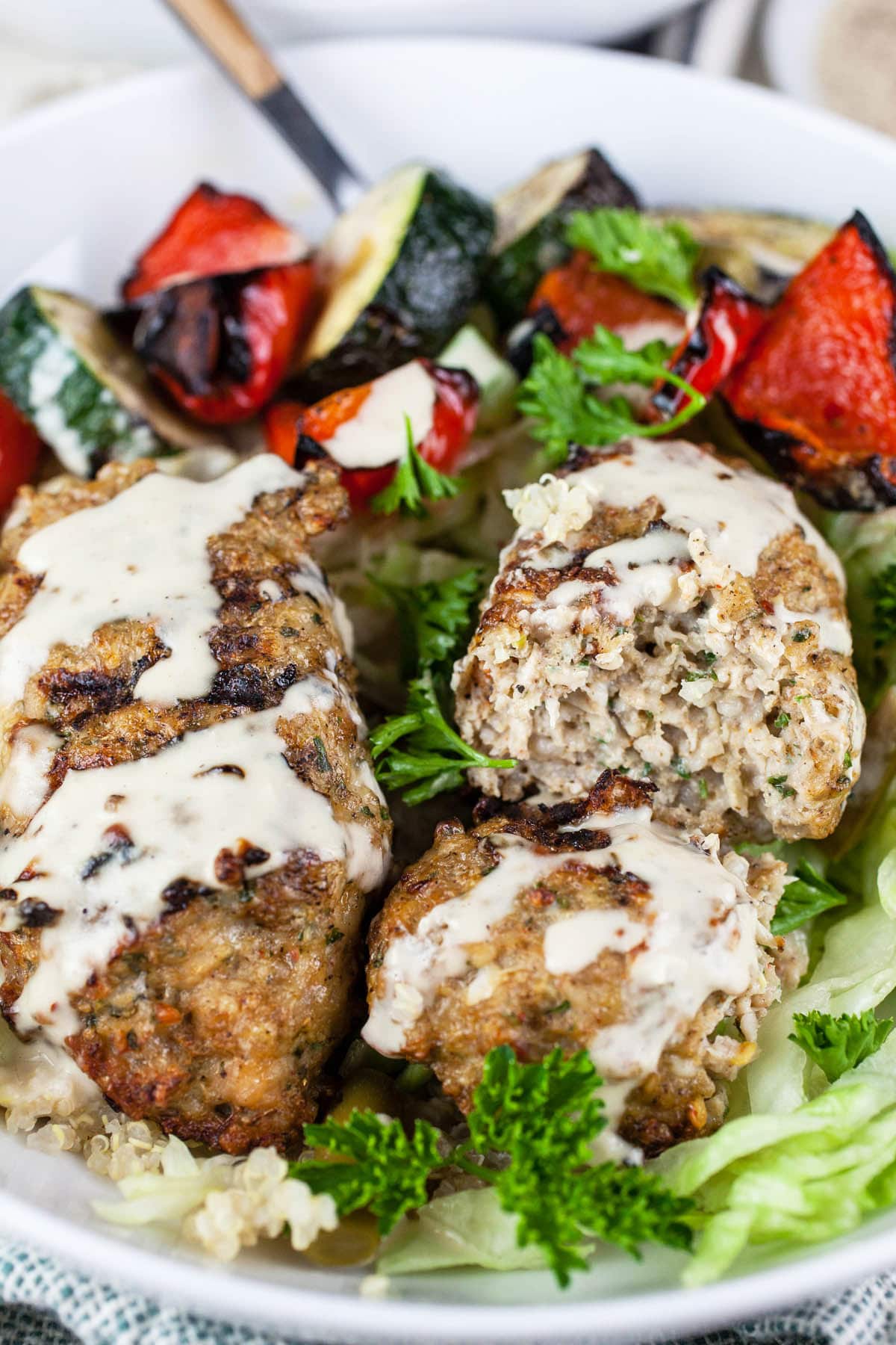 Chicken kofta with grilled vegetables and tahini sauce over quinoa in white bowl.