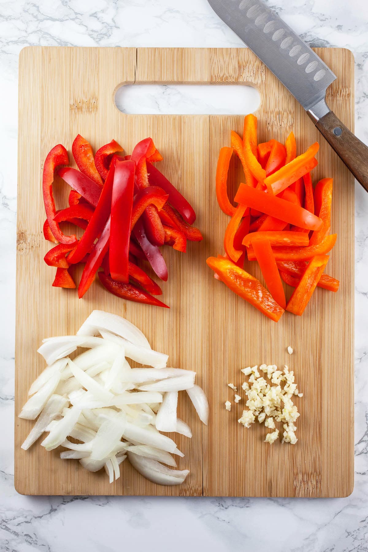 Minced garlic and sliced onions and red and orange bell peppers on wooden cutting board with knife.