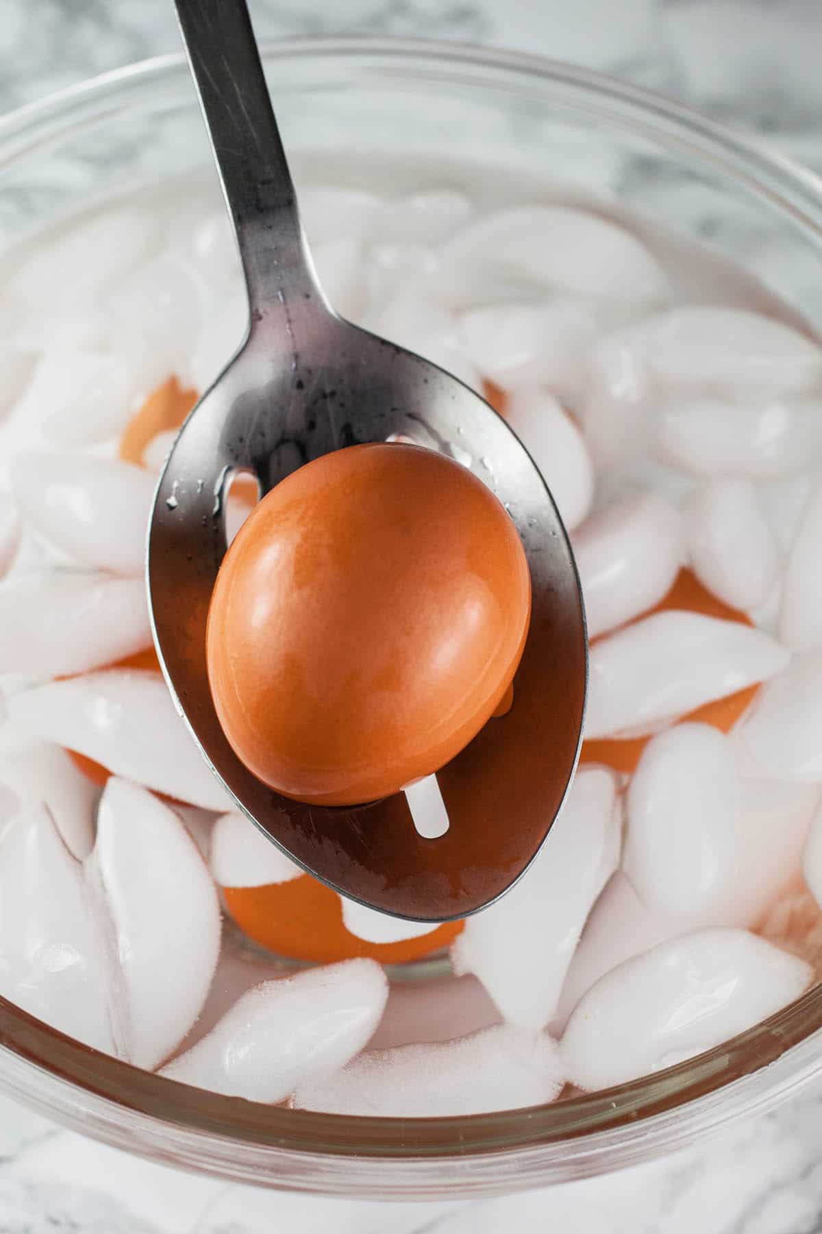Hard boiled egg on slotted spoon lifted from bowl of ice water.