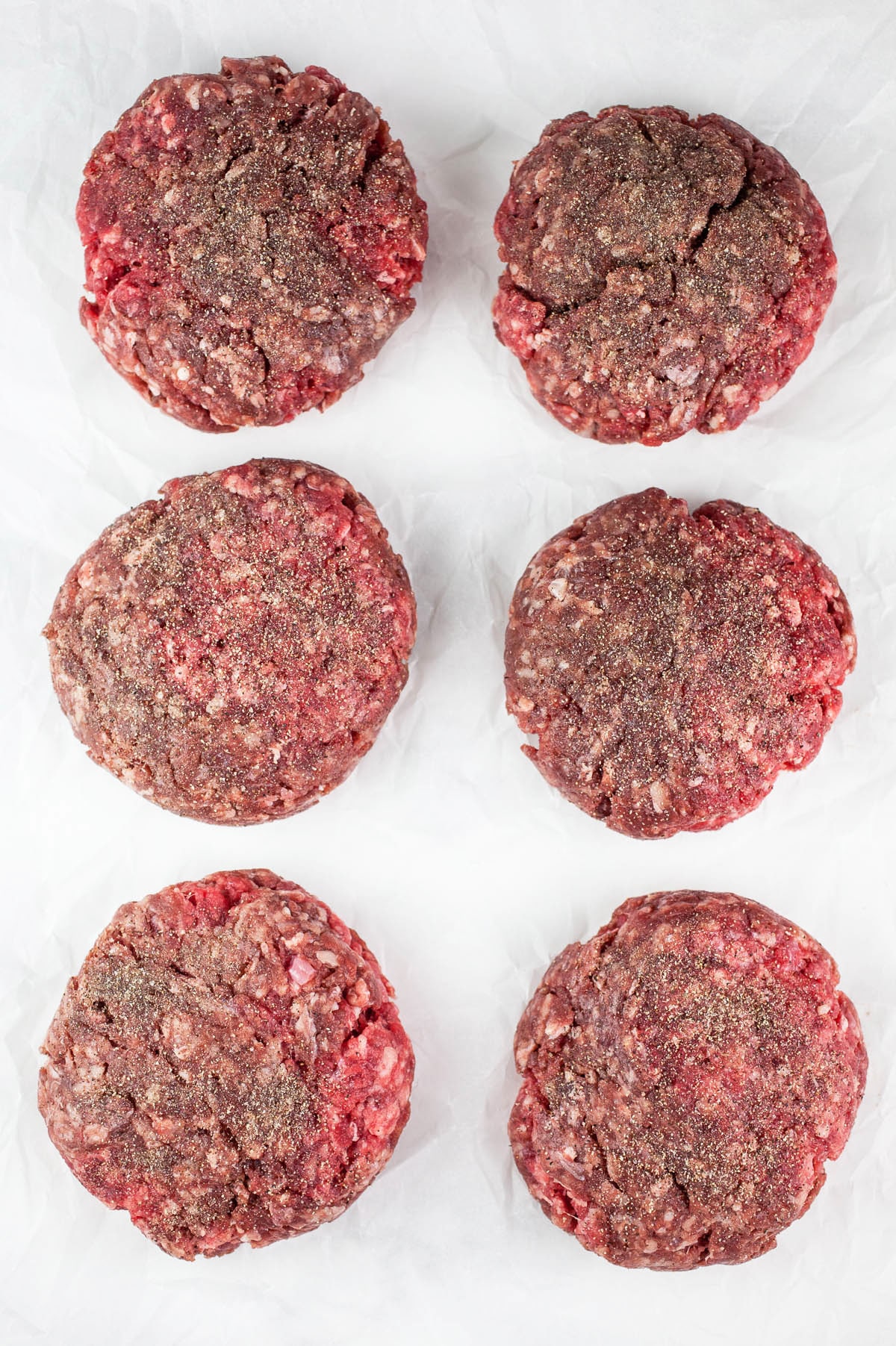 Raw ground beef patties with seasoning on white surface.