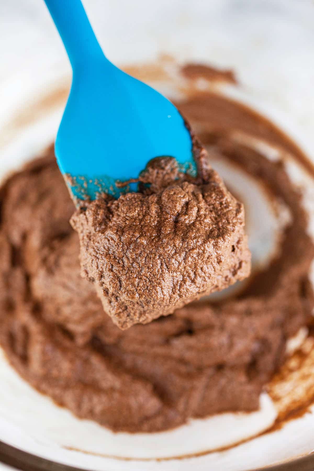 Chocolate frosting on blue spatula lifted from small glass bowl.