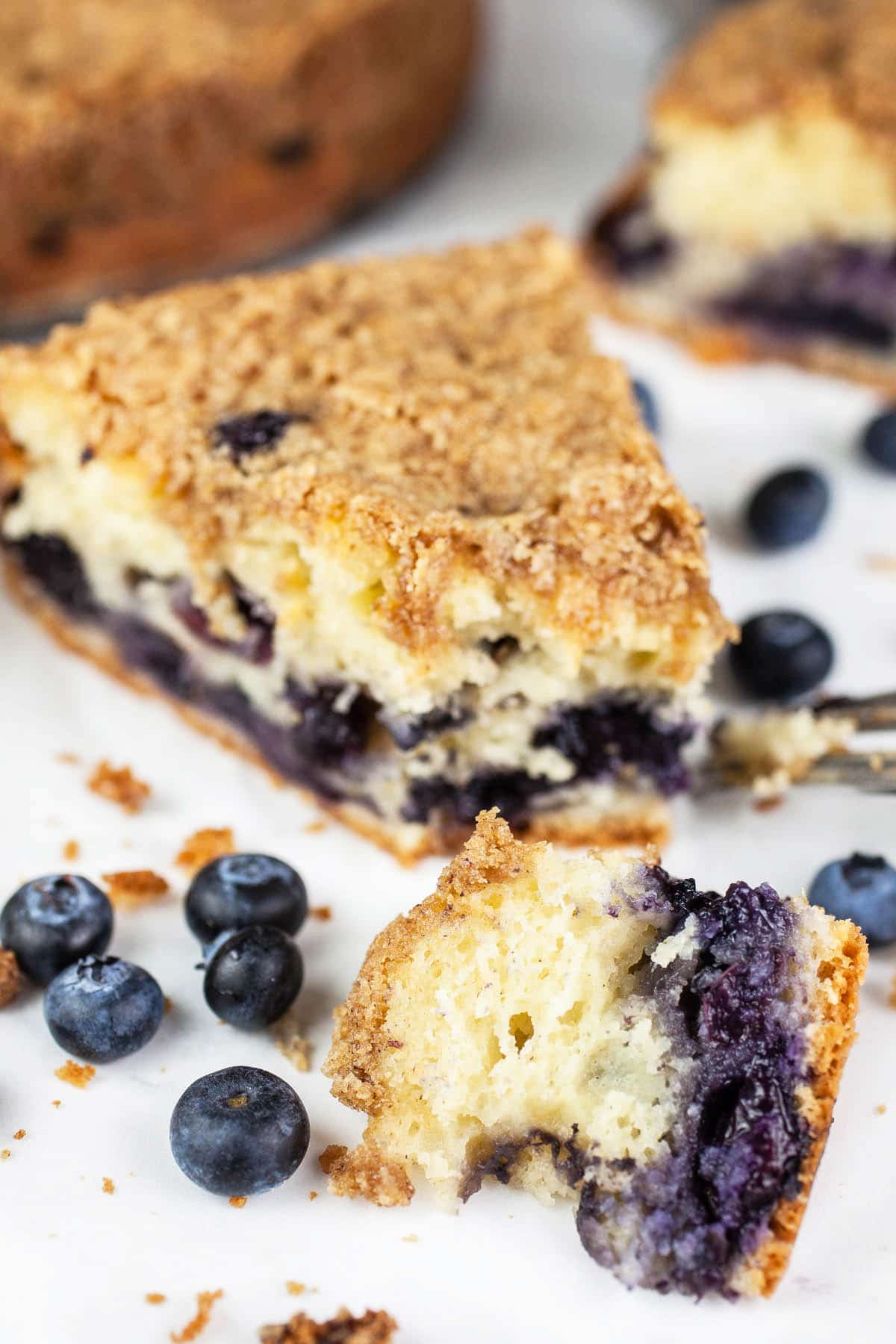Piece of blueberry coffee cake on white surface with fresh blueberries.