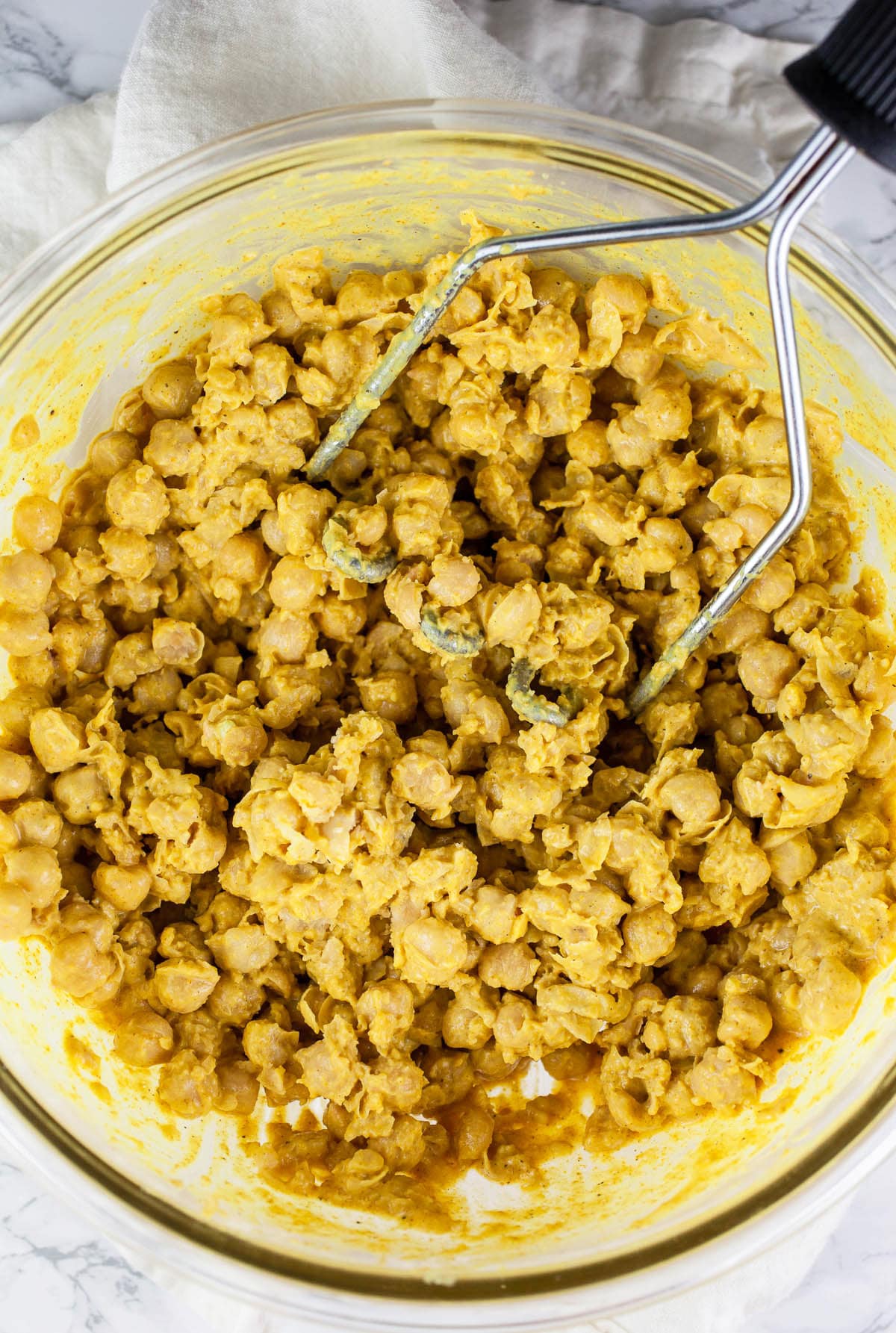 Mashed curried chickpeas in glass bowl with potato masher.