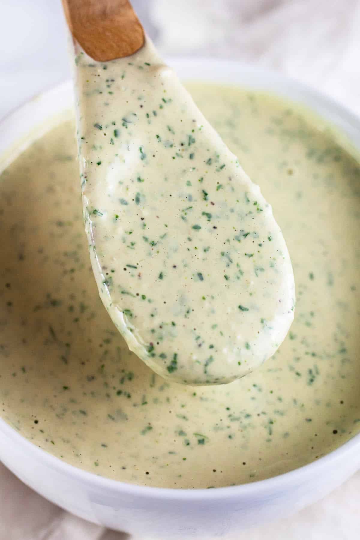 Wooden spoonful of herb tahini dressing lifted from small white bowl.