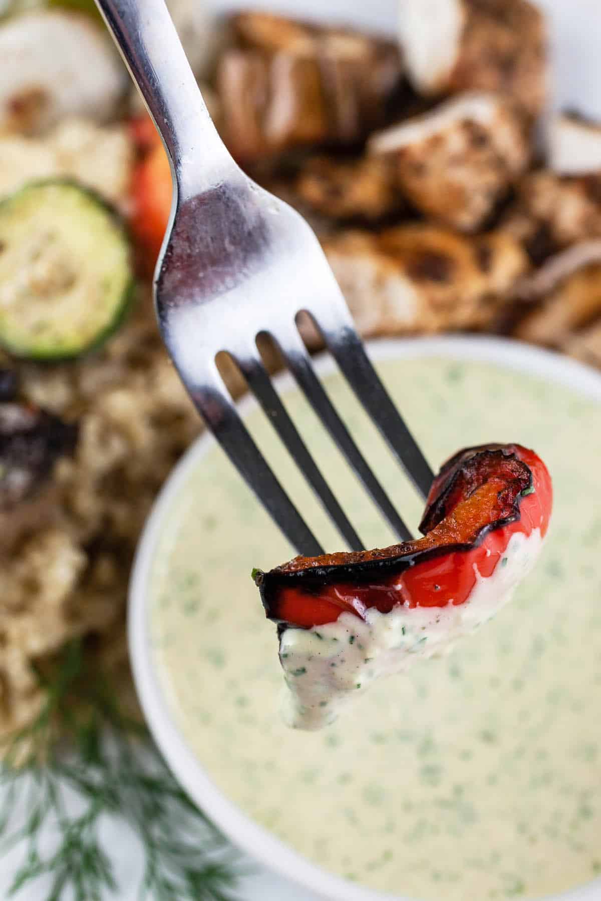 Grilled red pepper on fork dipped into small bowl of sauce.