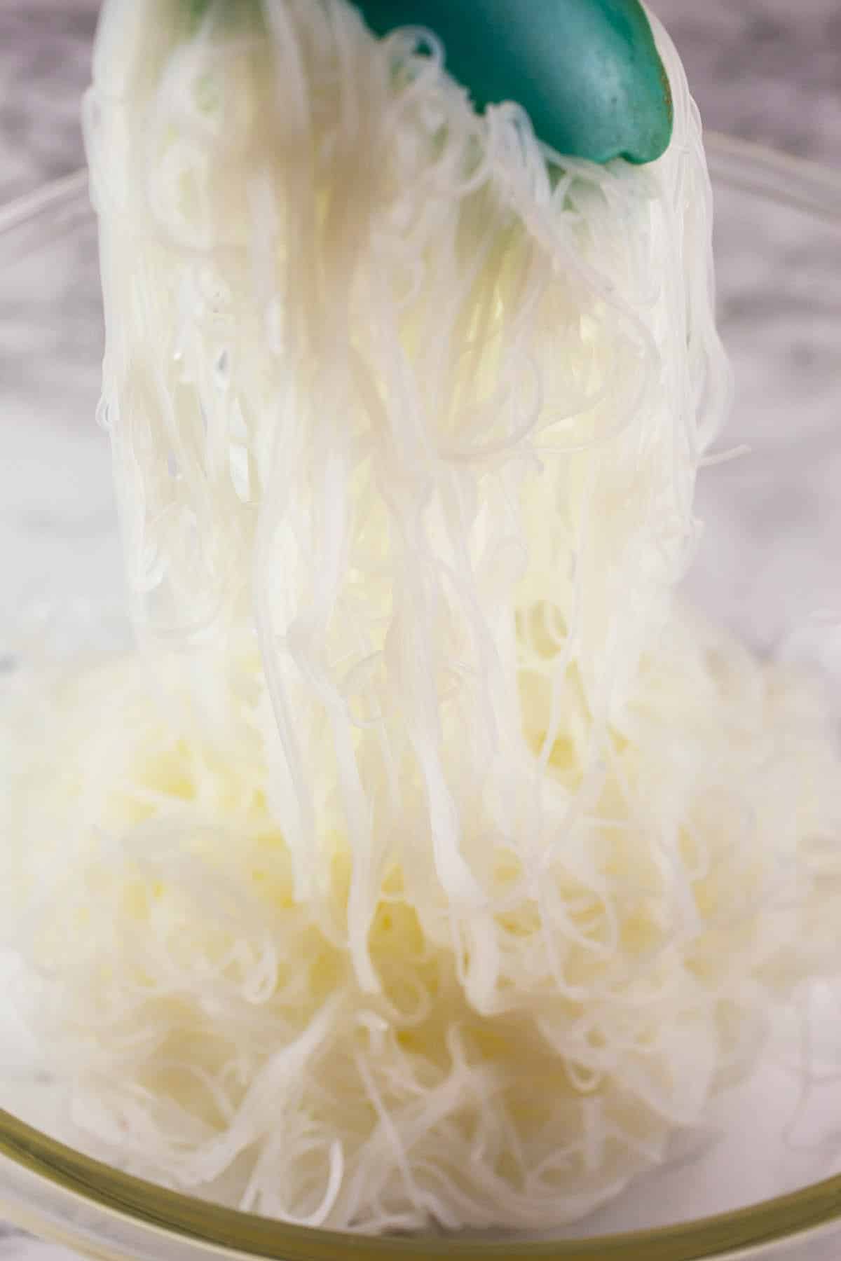 Vermicelli rice noodles lifted from large glass bowl with tongs.