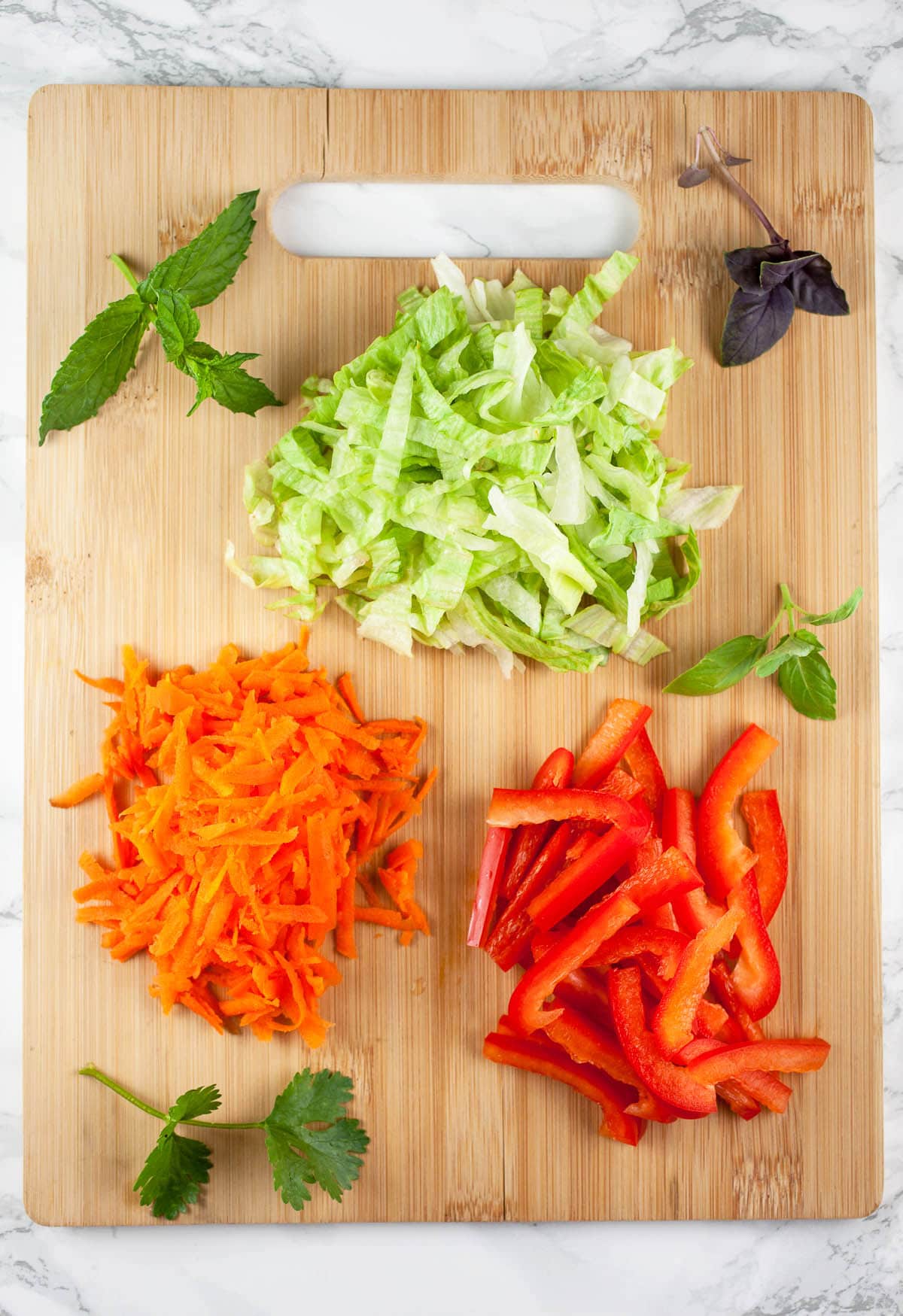 Shredded carrots, lettuce, red bell pepper, cilantro, basil, and mint on wooden cutting board.