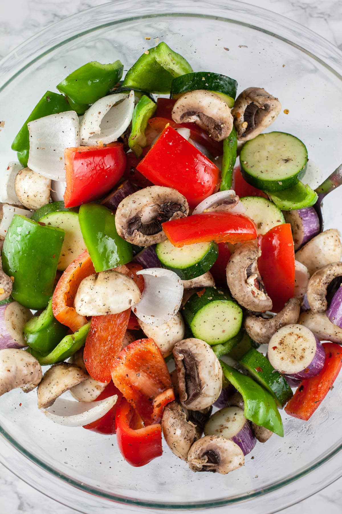 Chopped vegetables tossed in olive oil and spices in large glass bowl.