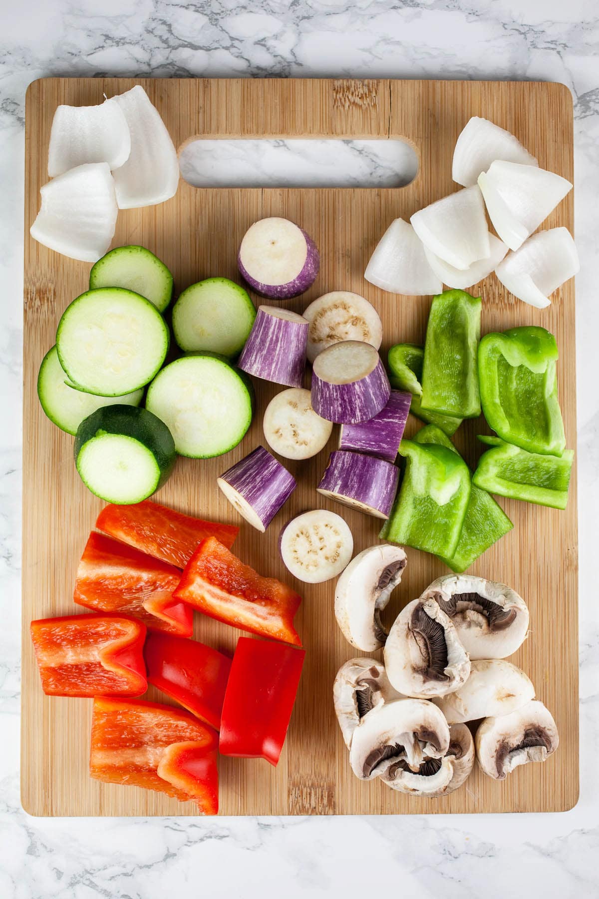 Red and green bell peppers, eggplant, mushrooms, zucchini, and onions on wooden cutting board.
