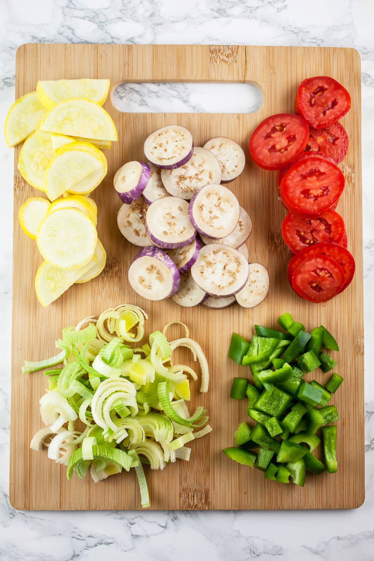 Chopped leeks, summer squash, eggplant, tomatoes, and green bell peppers on wooden cutting board.