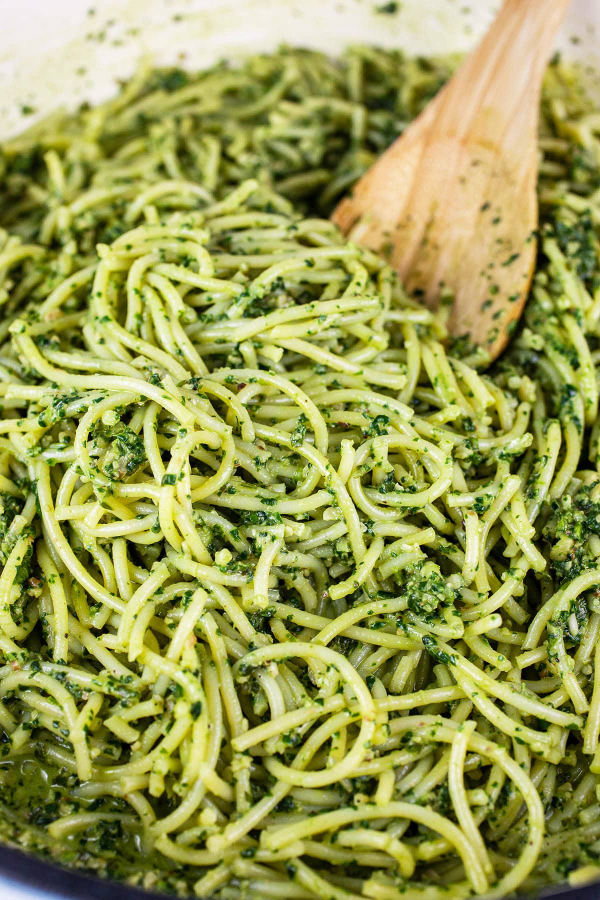 Spaghetti noodles tossed in pesto sauce with wooden spoon in skillet.