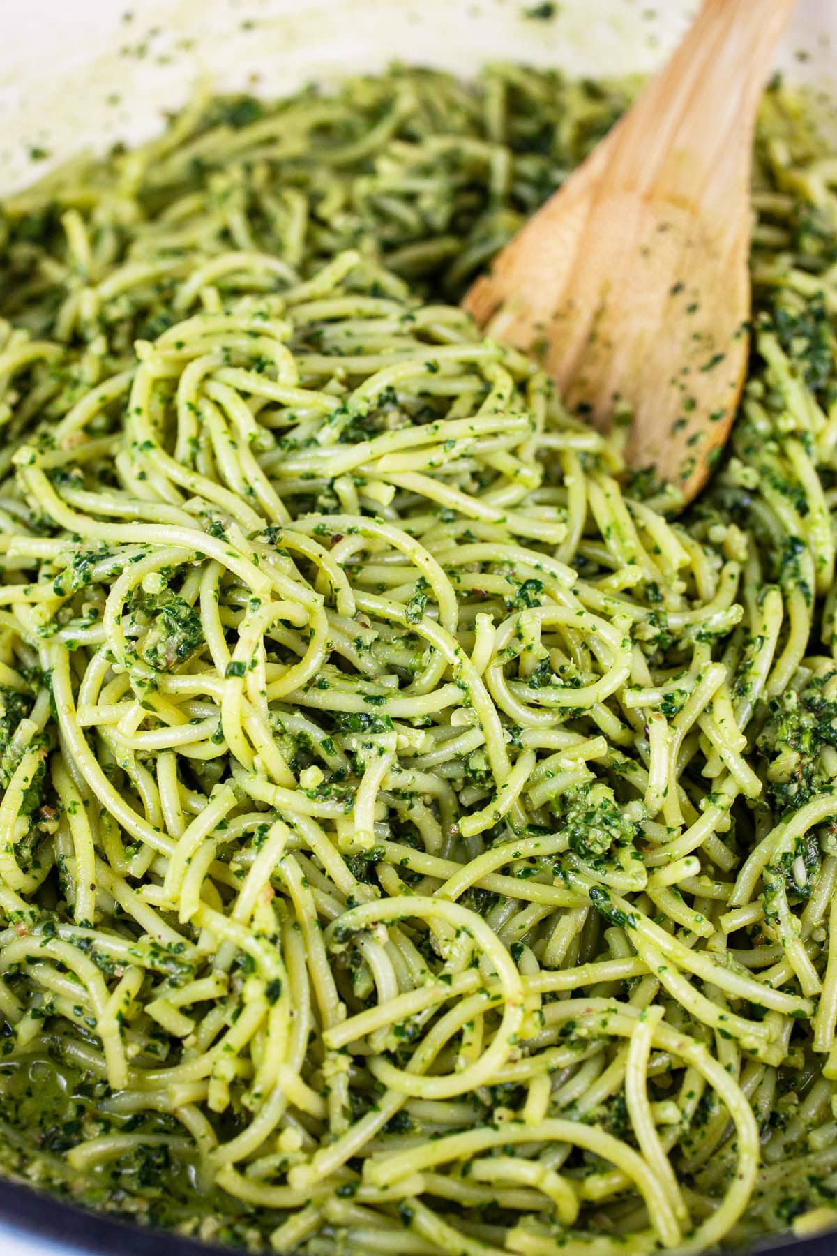 Spaghetti noodles tossed in pesto sauce with wooden spoon in skillet.