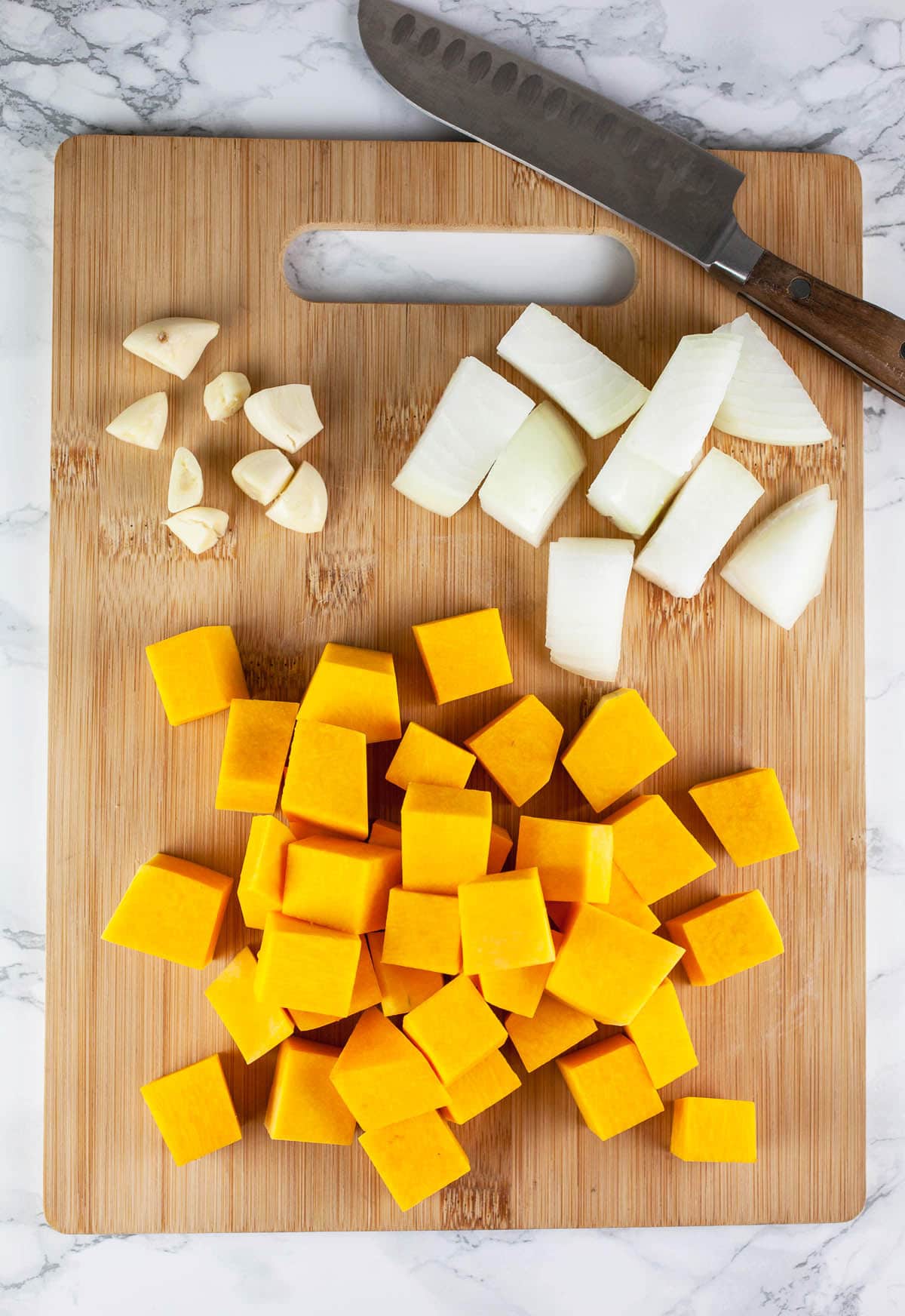 Chopped butternut squash, garlic, and onions on wooden cutting board with knife.