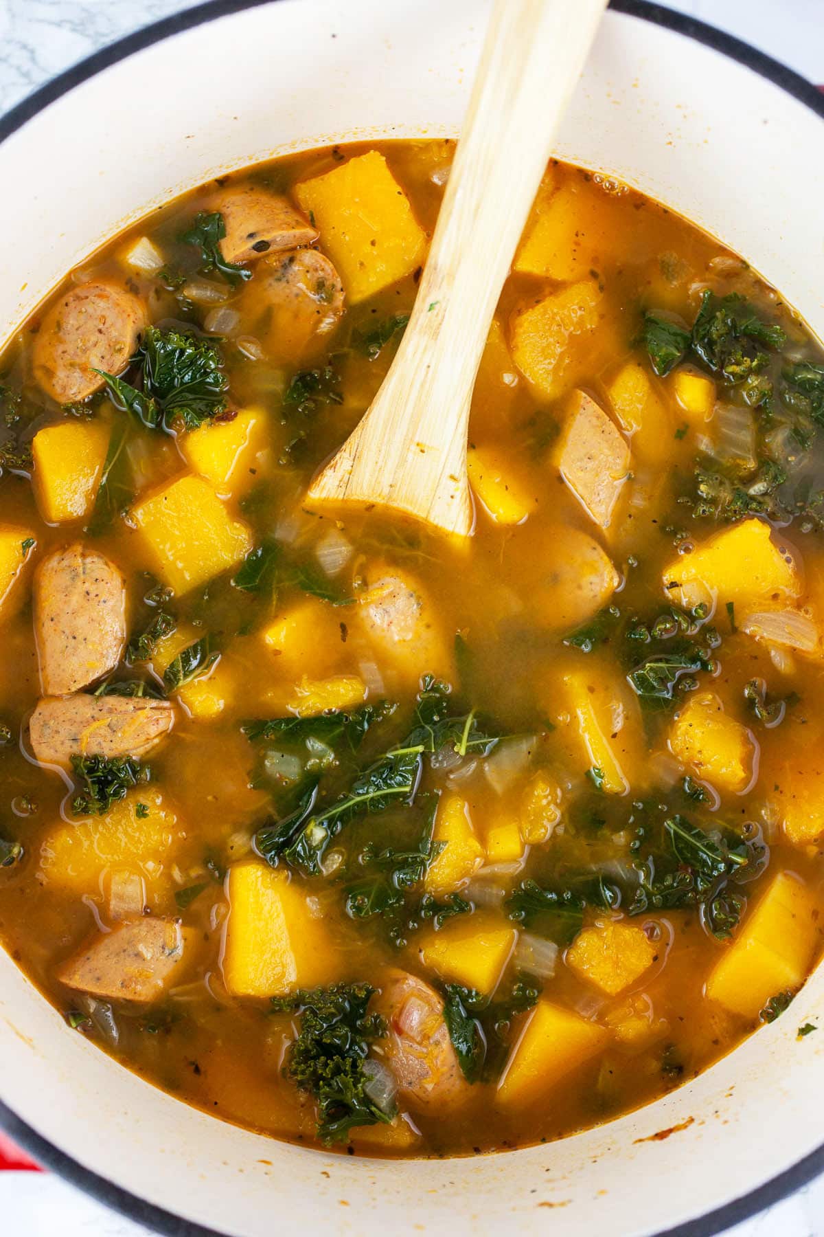 Chunky squash, sausage, and kale soup in cast iron Dutch oven with wooden spoon.