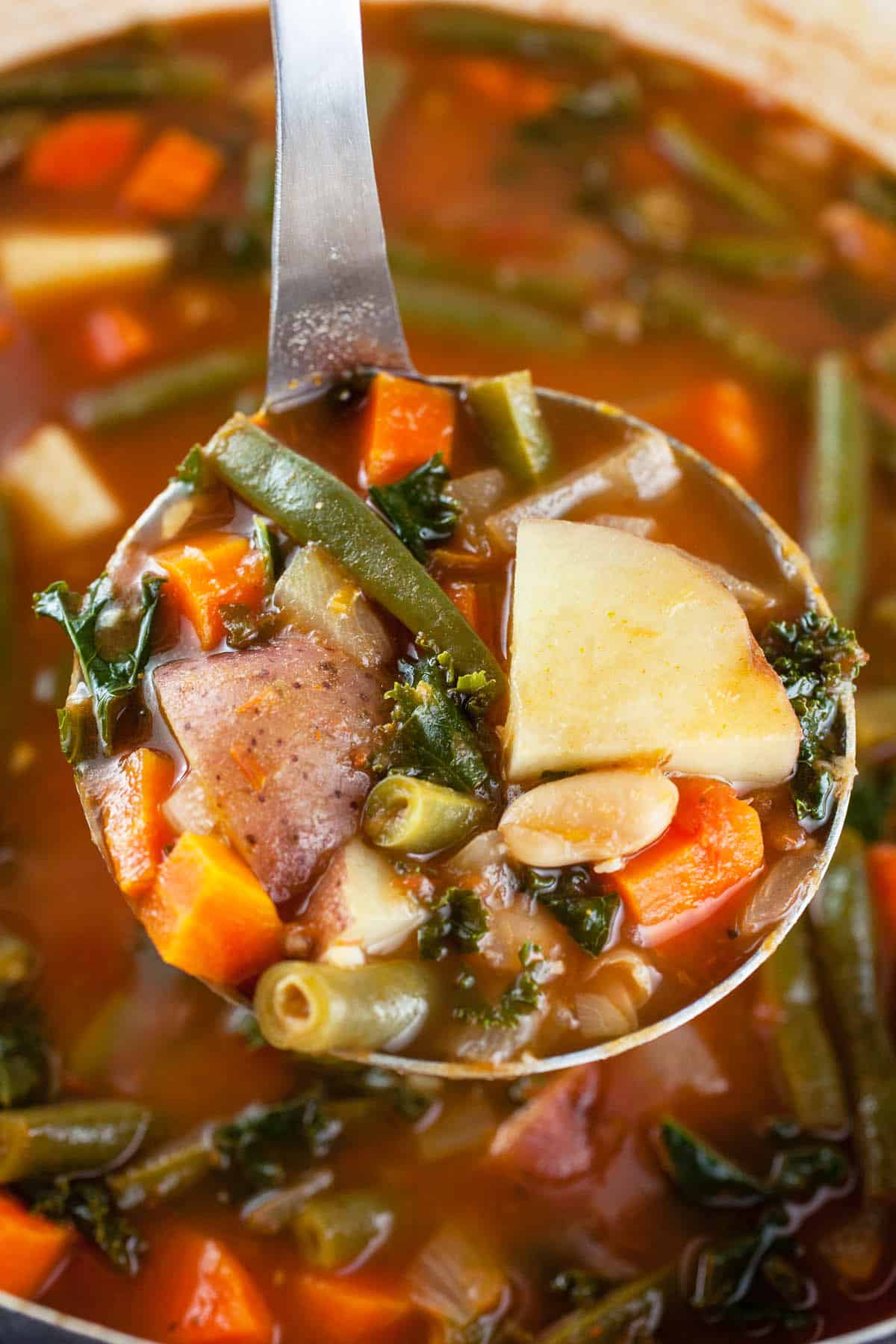 Scoop of vegetable soup in metal ladle lifted from Dutch oven.