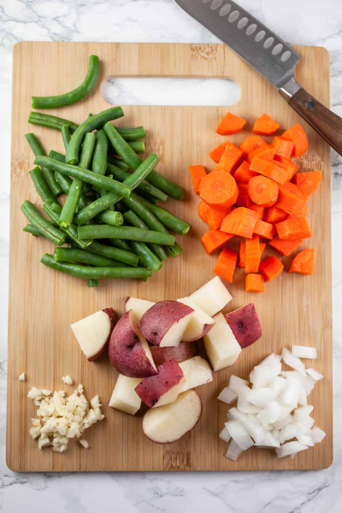 Minced garlic, onions, diced red potatoes, green beans, and carrots on wooden cutting board with knife.