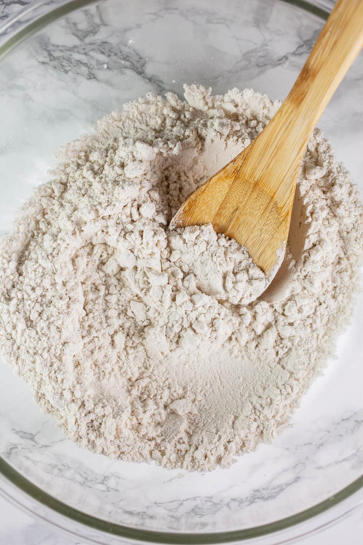 Dry ingredients in large glass bowl with wooden spoon.