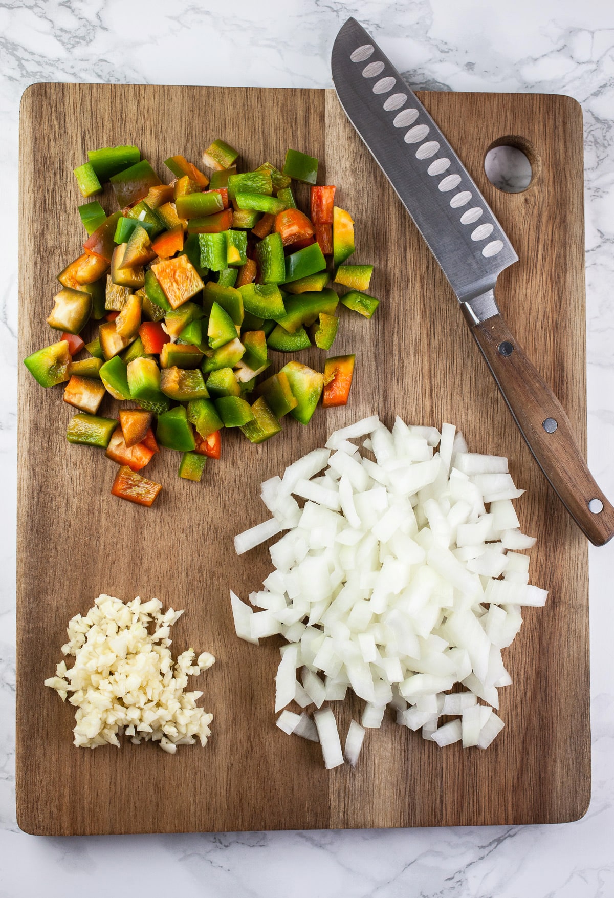 Minced garlic, onions, and bell peppers on wooden cutting board with knife.